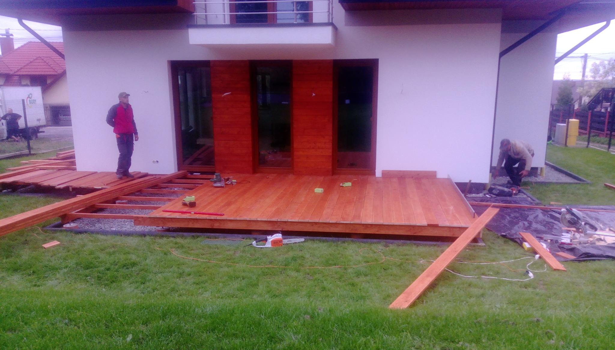 Budowa drewnianego tarasu przy nowoczesnym domu z drewnianą elewacją, widoczni dwaj pracownicy, jeden w czerwonej kurtce stoi na konstrukcji, drugi pracuje na ziemi, narzędzia rozłożone na trawie.