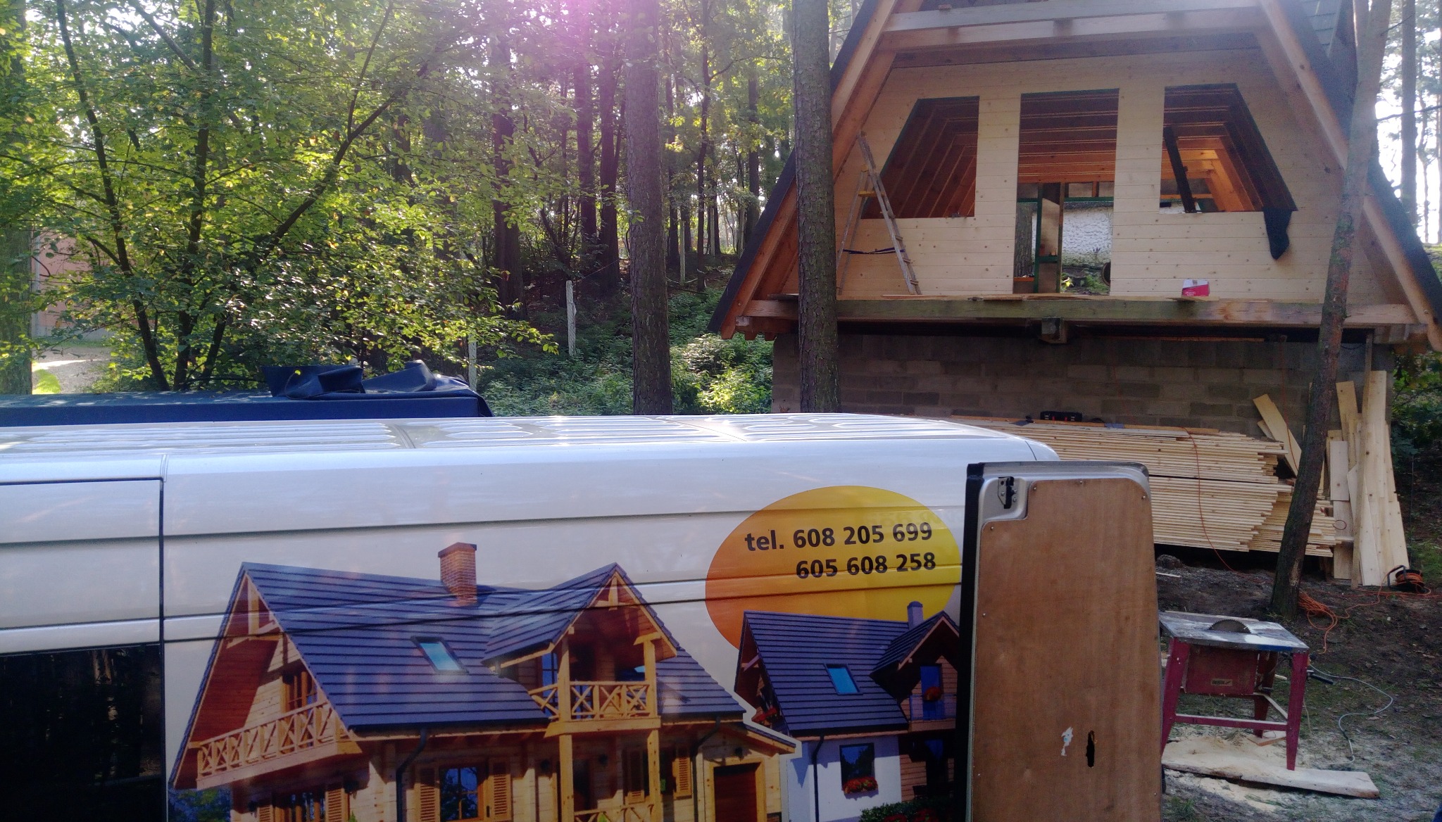 Wznoszona konstrukcja drewniana w lesie, widoczny fragment samochodu firmowego z numerami telefonów i grafiką domu, stos desek i piła stołowa.
