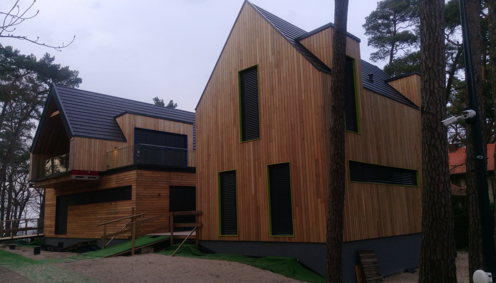 Nowoczesny dom z drewnianą elewacją, czarnymi oknami i dachem, widoczny system monitoringu, otoczony drzewami.