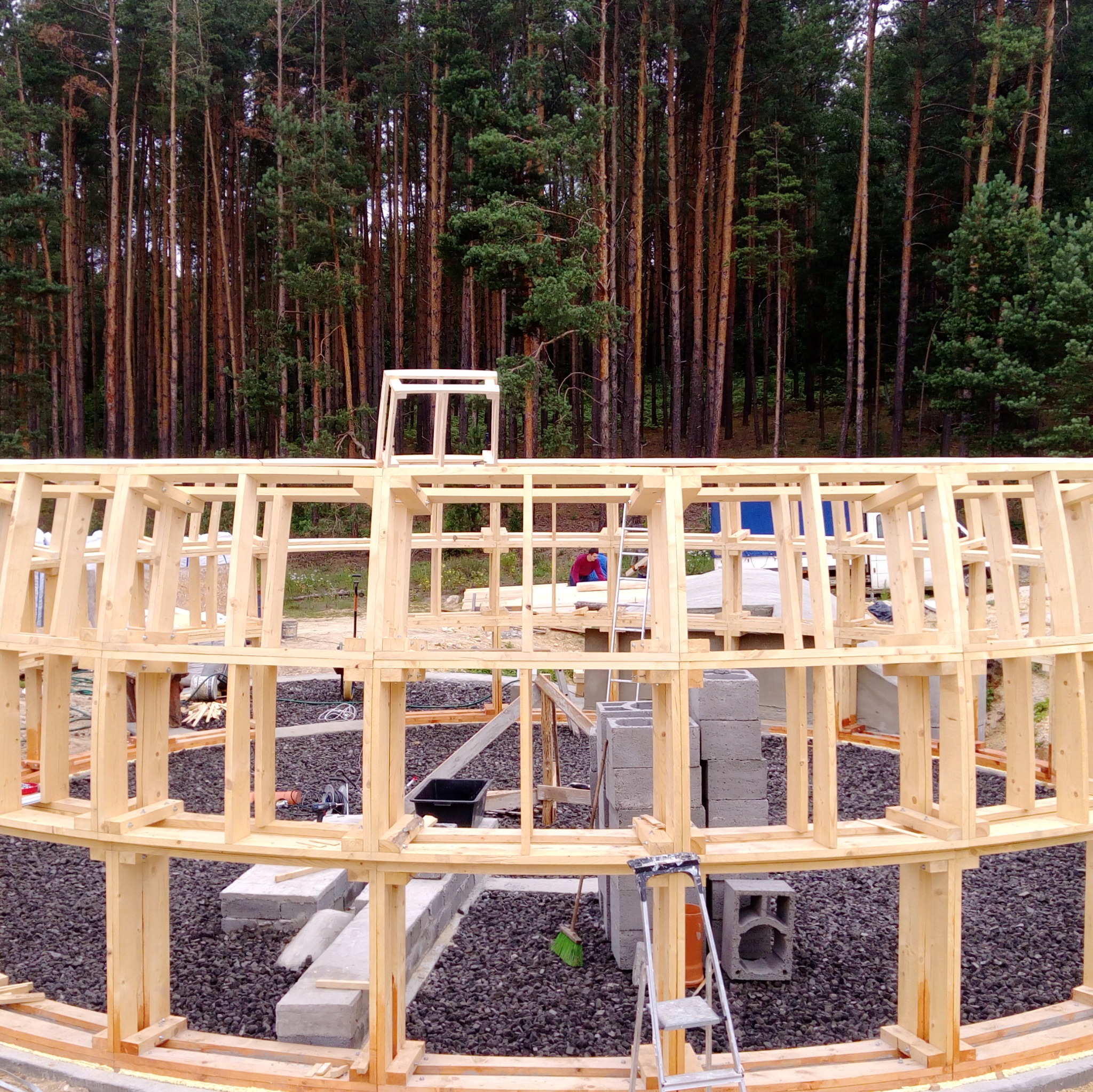 Konstrukcja szkieletowa budynku w trakcie budowy, widoczna na tle lasu, z detalami takimi jak drabina, bloczki betonowe i czarne kamienie na ziemi.