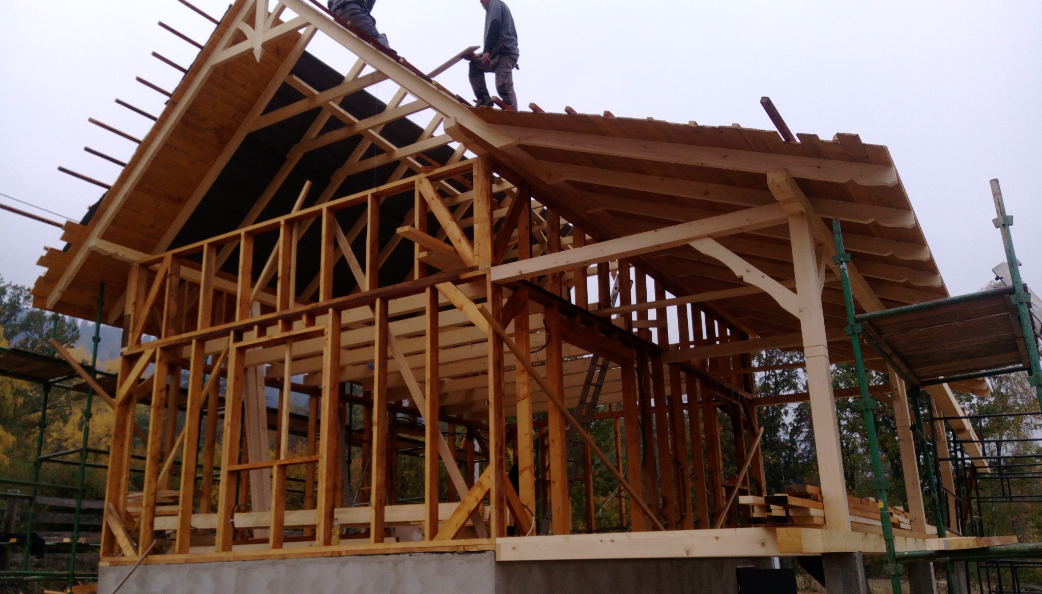 Wznoszenie drewnianej konstrukcji domu szkieletowego z widocznymi elementami więźby dachowej i pracownikami na dachu, uchwycone w pochmurny dzień.