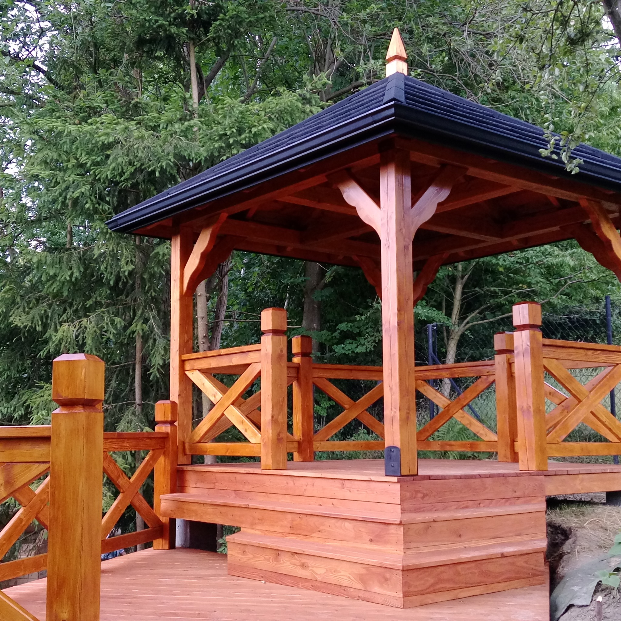 Drewniana altana z czarnym dachem, otoczona zielenią drzew, z barierkami w kształcie krzyżyków i schodkami prowadzącymi na podest.