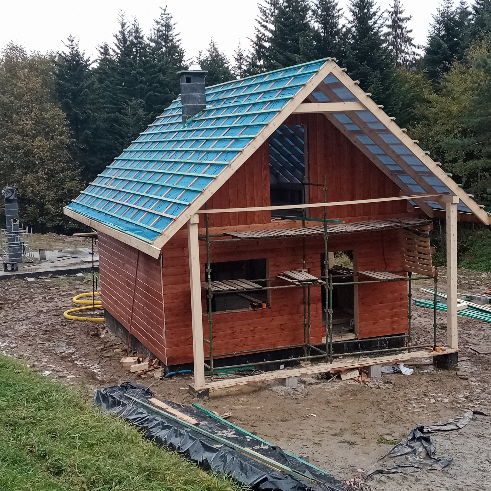 Niedokończony, drewniany budynek z niebieską membraną dachową, otoczony rusztowaniem i błotnistym terenem, z lasem w tle.