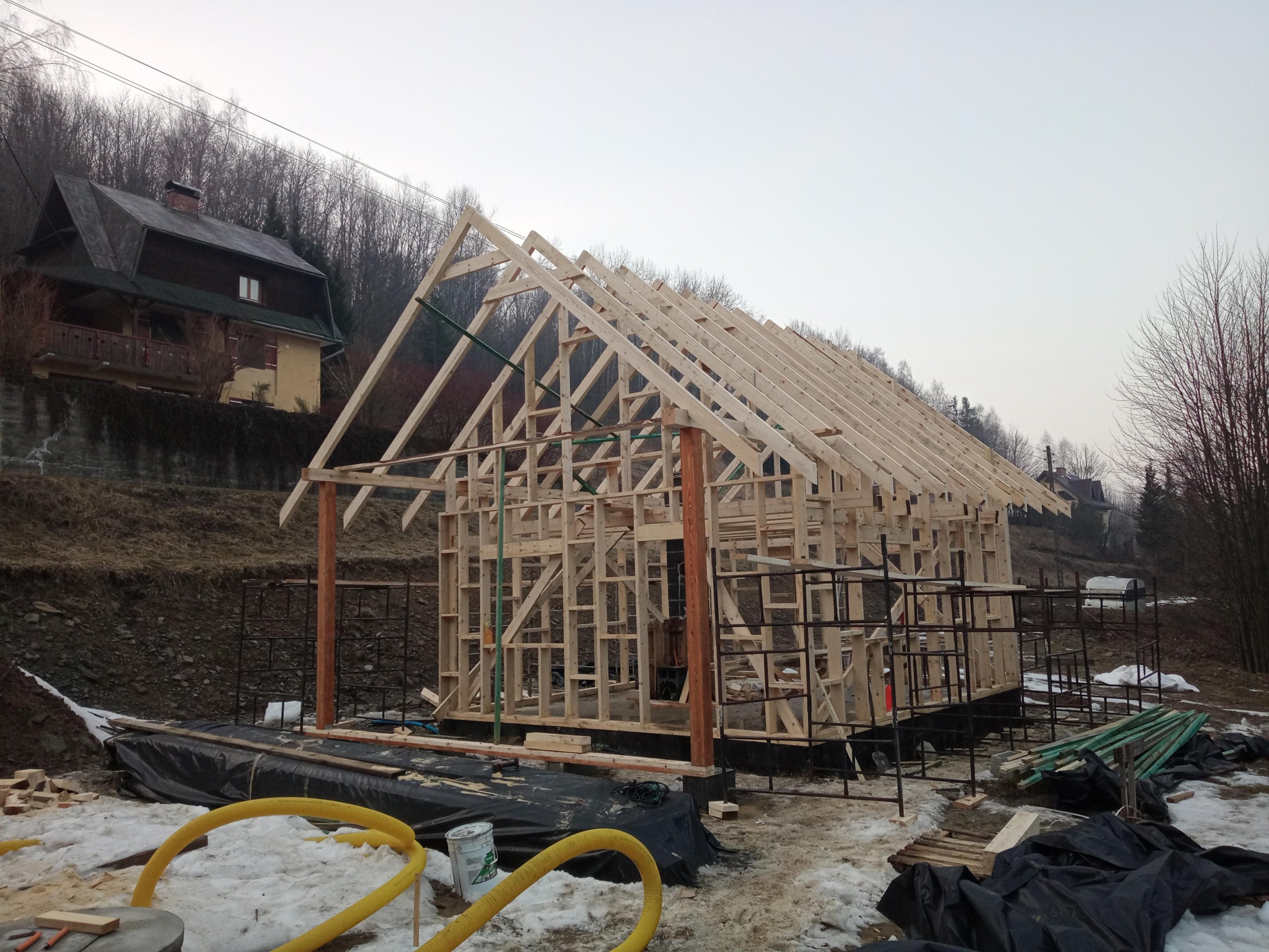 Surowy drewniany szkielet powstającego domu jednorodzinnego na tle zalesionego wzgórza i istniejącego budynku.