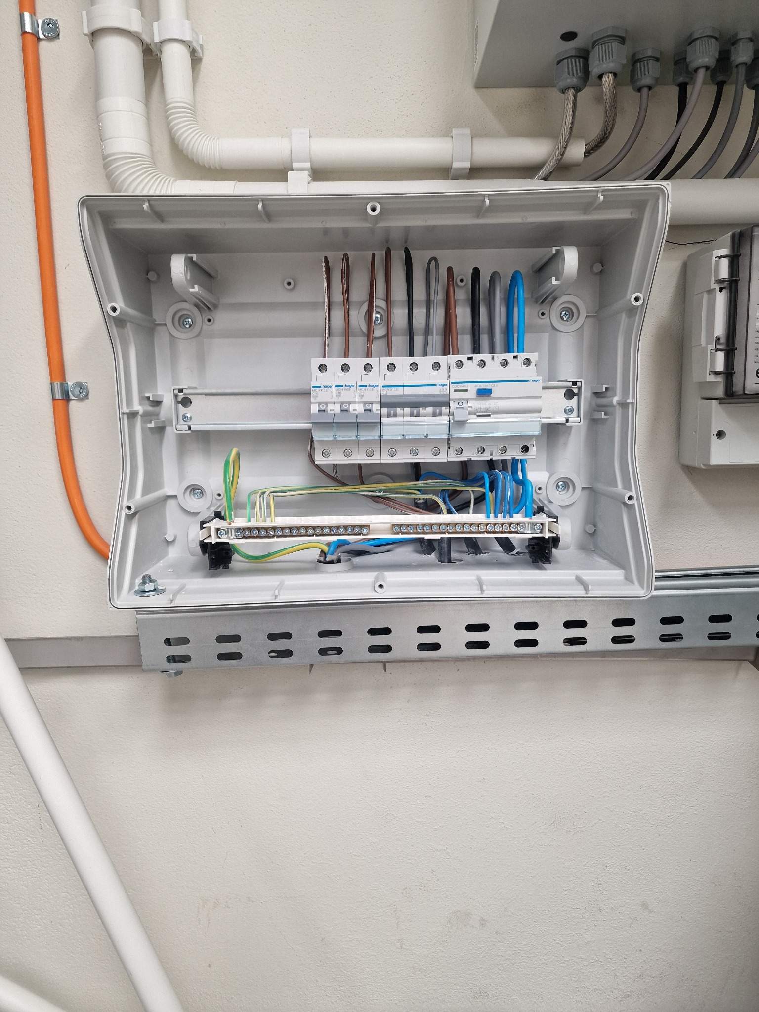 Otwarta skrzynka elektryczna z widocznymi przewodami, bezpiecznikami i listwą zaciskową, zamontowana na ścianie z rurami instalacyjnymi.