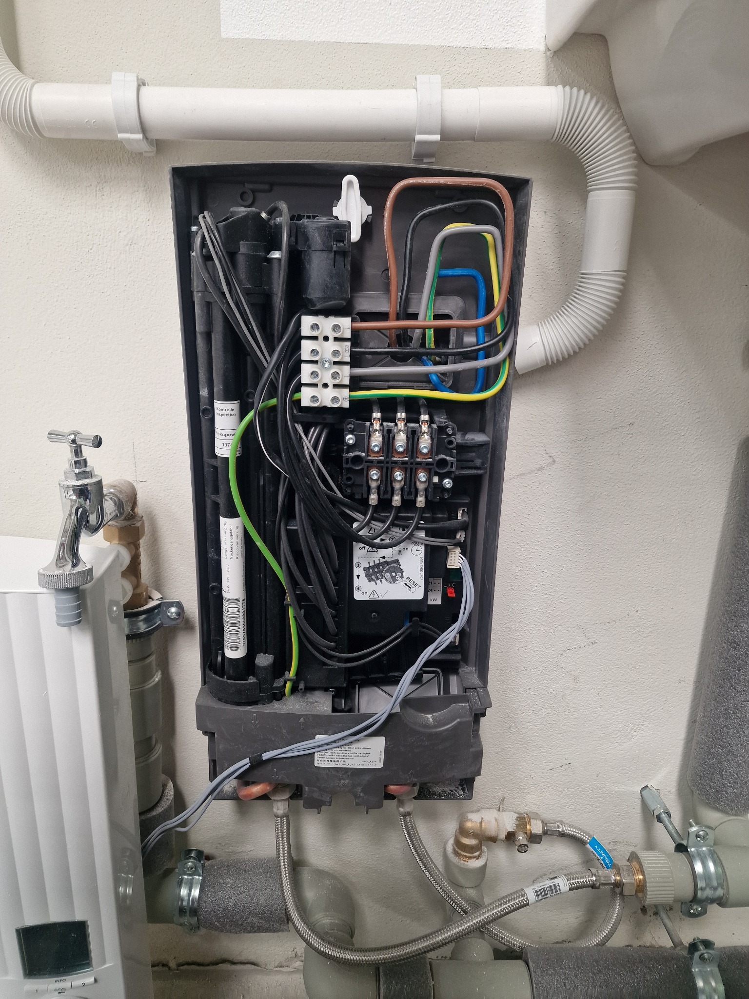 Otwarta obudowa urządzenia elektrycznego na ścianie z widocznymi przewodami, złączami i elementami instalacji hydraulicznej. Widoczne rury i zawór.