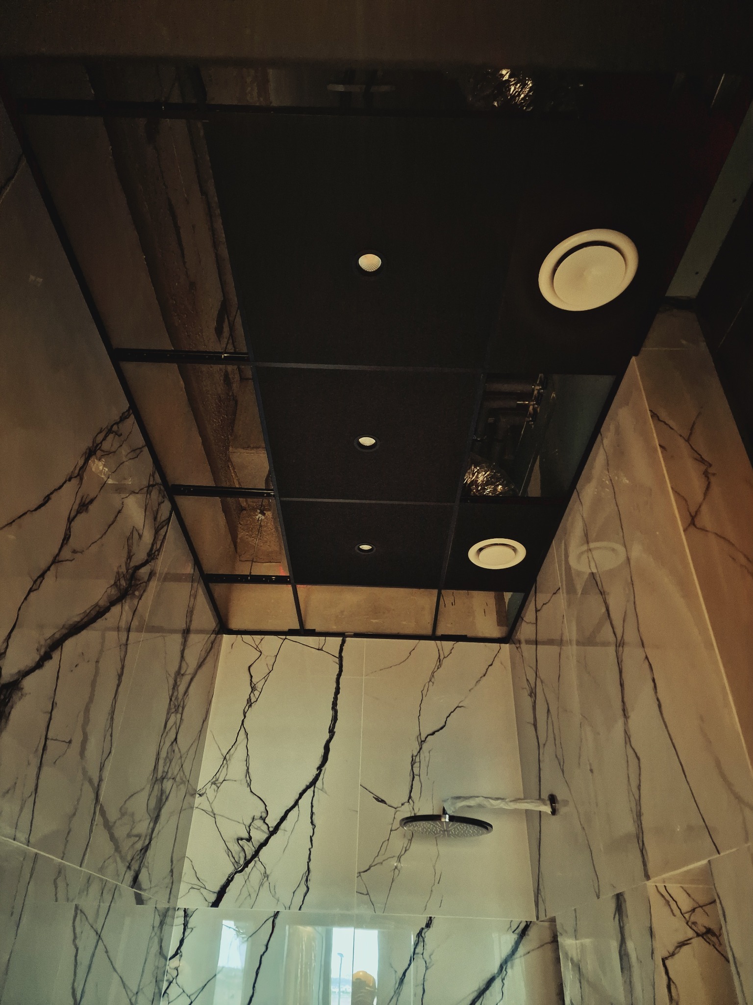 Nowoczesna łazienka z marmurowymi płytkami, czarnym sufitem podwieszanym z oświetleniem punktowym i wentylacją. Widok z dołu na kabinę prysznicową.