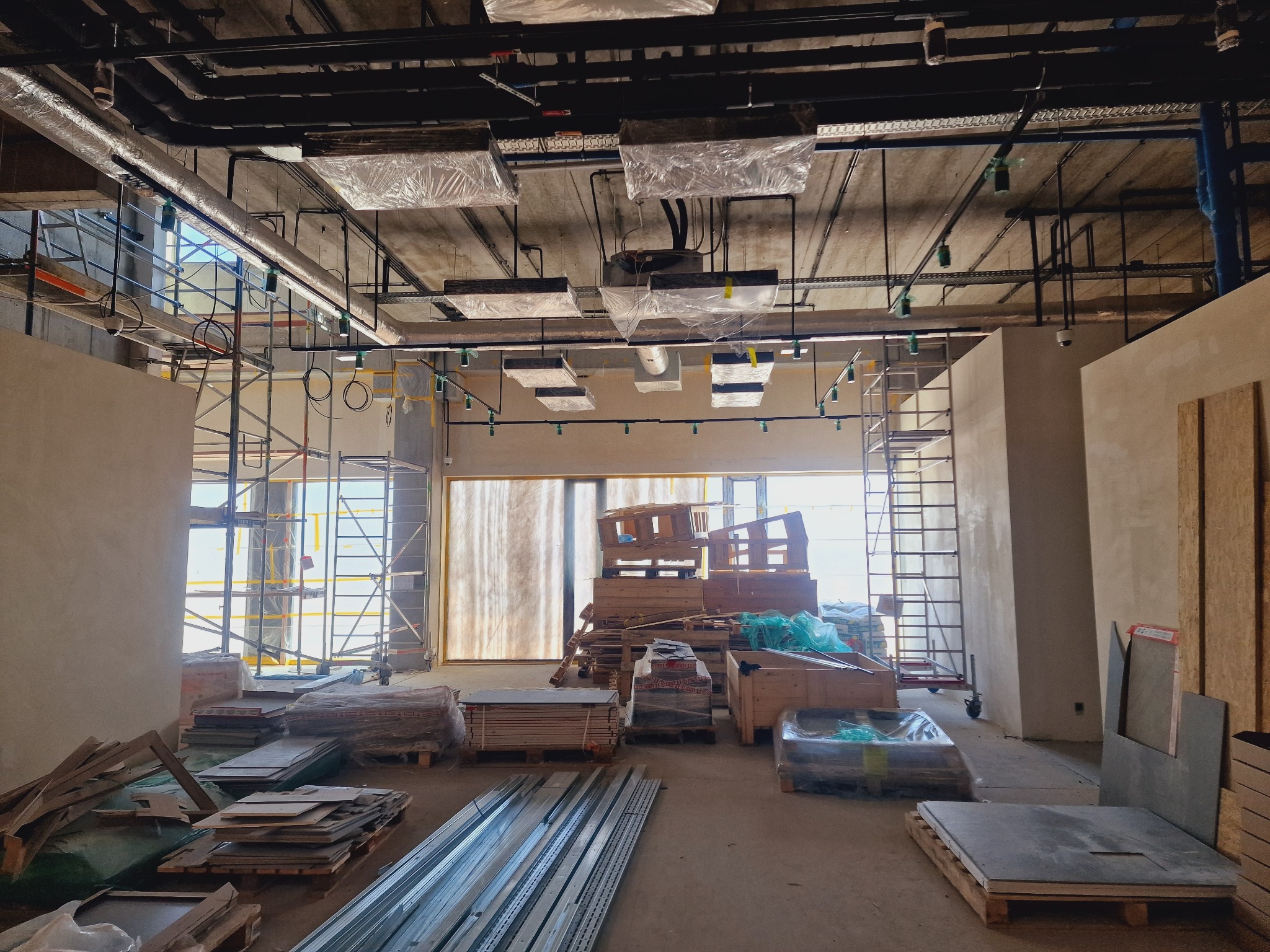 Wnętrze w trakcie prac instalacyjnych: widoczne rury, kable pod sufitem, rusztowania i materiały budowlane, oświetlenie tymczasowe. Surowy stan wykończenia.