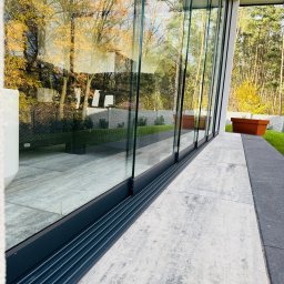 SEBAMET SEBASTIAN GRACZ - Przesuwne szklane drzwi tarasowe z czarną ramą, widok na jesienny ogród z betonowym ogrodzeniem i donicami.