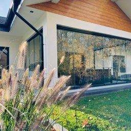SEBAMET SEBASTIAN GRACZ - Nowoczesny dom z panoramicznym oknem przesuwnym, odbijającym jesienny krajobraz, widoczne trawy ozdobne na pierwszym planie.