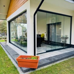 SEBAMET SEBASTIAN GRACZ - Nowoczesne, przesuwne szklane drzwi tarasowe z czarną ramą, widok z zewnątrz, z psem rasy Jack Russell Terrier stojącym na tarasie i odbiciem drugiego psa w szybie.