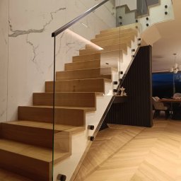 SEBAMET SEBASTIAN GRACZ - Nowoczesne schody drewniane z balustradą szklaną, mocowaną punktowo, oświetlone listwą LED, na tle ściany imitującej marmur i parkietu w jodełkę.