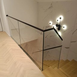 SEBAMET SEBASTIAN GRACZ - Szklana balustrada z czarnymi elementami wykończeniowymi zamontowana przy schodach z drewnianymi stopniami, oświetlona nowoczesną lampą na marmurowej ścianie.