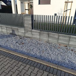 SEBAMET SEBASTIAN GRACZ - Nowoczesne ogrodzenie z szarych, betonowych bloczków i pionowych, metalowych przęseł, oddzielające posesję od chodnika wyłożonego kostką brukową, z ozdobnym wysypaniem kamykami.