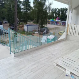 Turkusowa balustrada metalowa i szklana na tarasie z widokiem na plac zabaw i okoliczną zieleń.