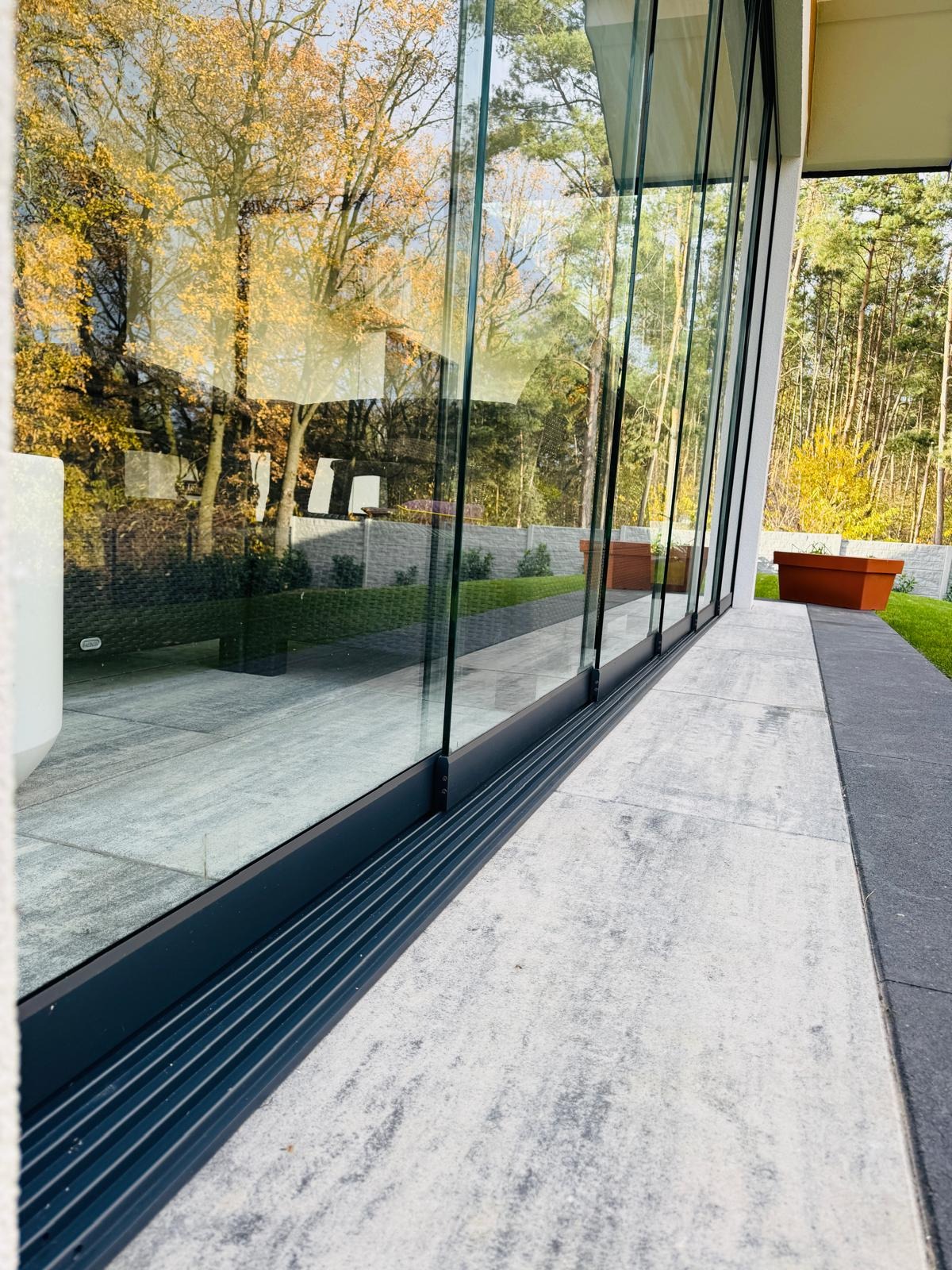 Przesuwne szklane drzwi tarasowe z czarną ramą, widok na jesienny ogród z betonowym ogrodzeniem i donicami.