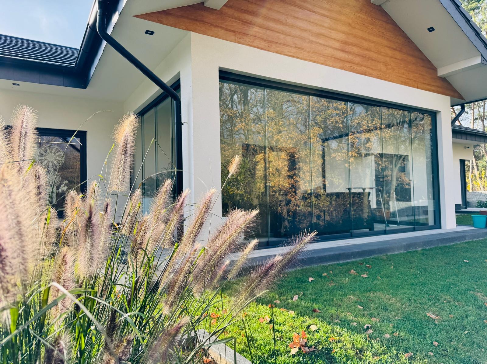 Nowoczesny dom z panoramicznym oknem przesuwnym, odbijającym jesienny krajobraz, widoczne trawy ozdobne na pierwszym planie.