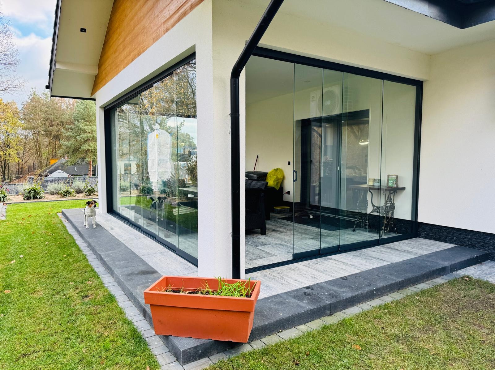 Nowoczesne, przesuwne szklane drzwi tarasowe z czarną ramą, widok z zewnątrz, z psem rasy Jack Russell Terrier stojącym na tarasie i odbiciem drugiego psa w szybie.