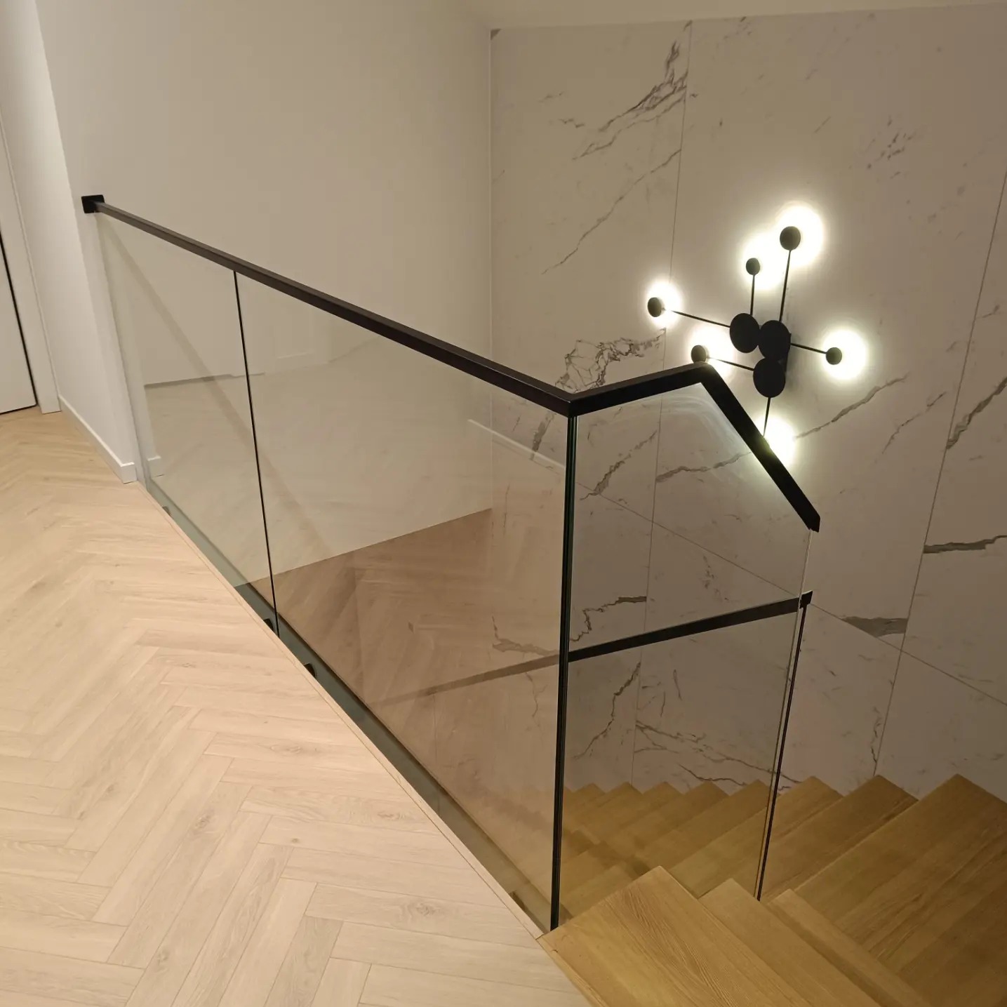Szklana balustrada z czarnymi elementami wykończeniowymi zamontowana przy schodach z drewnianymi stopniami, oświetlona nowoczesną lampą na marmurowej ścianie.