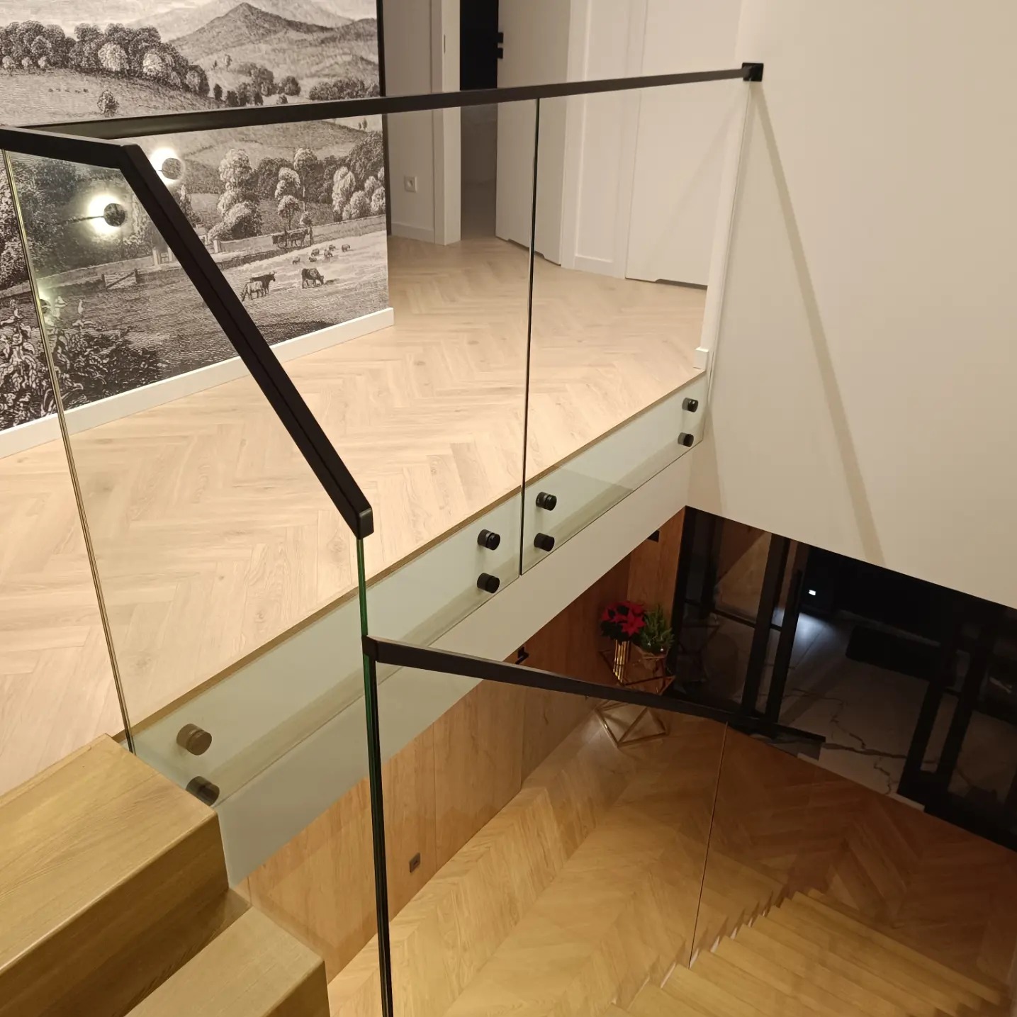 Szklana balustrada z czarnymi elementami mocującymi, widok z góry na drewniane schody i parkiet ułożony w jodełkę, w tle dekoracyjna tapeta z motywem wiejskim.