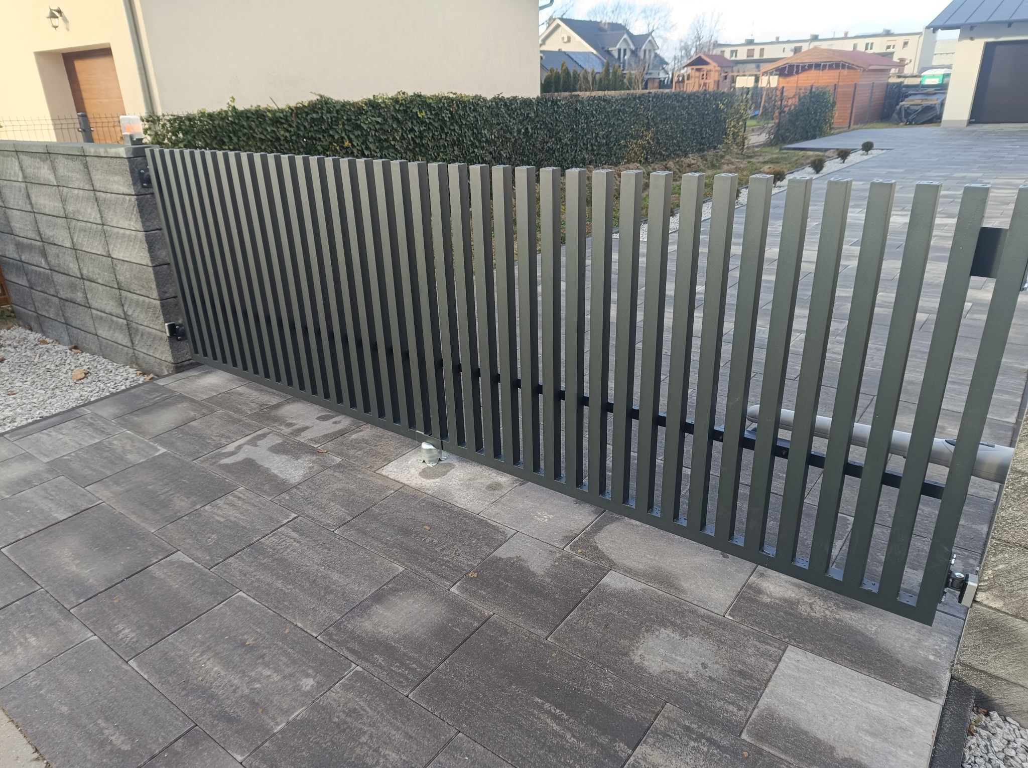Nowoczesna, szara brama wjazdowa z pionowych profili, widoczny mechanizm automatycznego otwierania, kostka brukowa w odcieniach szarości, żywopłot w tle.