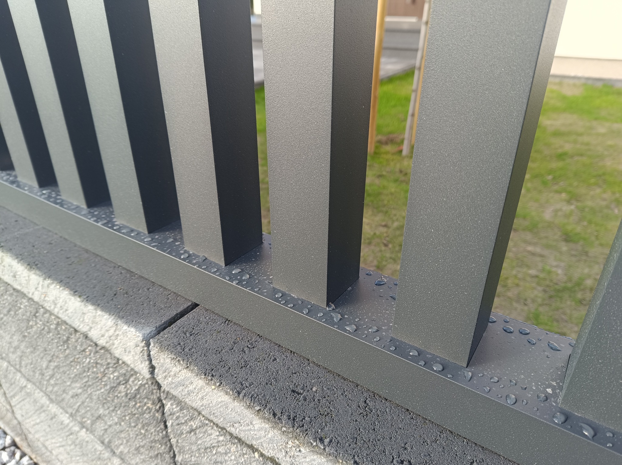 Fragment nowoczesnego ogrodzenia metalowego w kolorze grafitowym z kroplami wody na poziomych elementach, osadzonego na betonowej podmurówce.