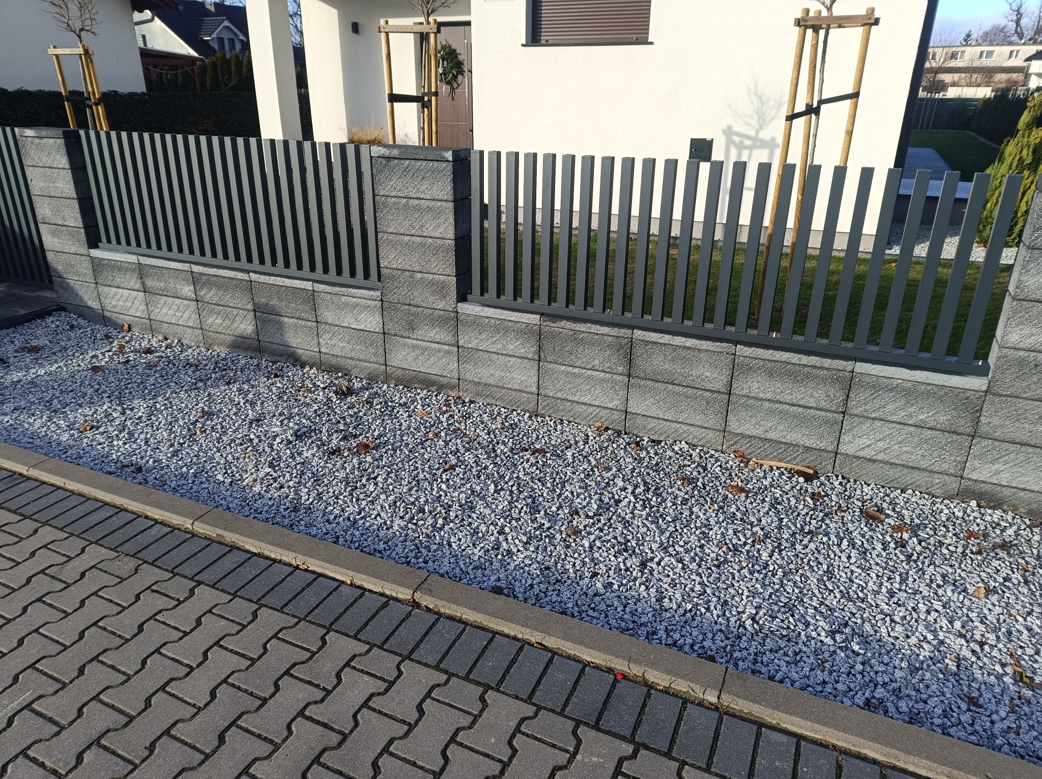 Nowoczesne ogrodzenie z szarych, betonowych bloczków i pionowych, metalowych przęseł, oddzielające posesję od chodnika wyłożonego kostką brukową, z ozdobnym wysypaniem kamykami.