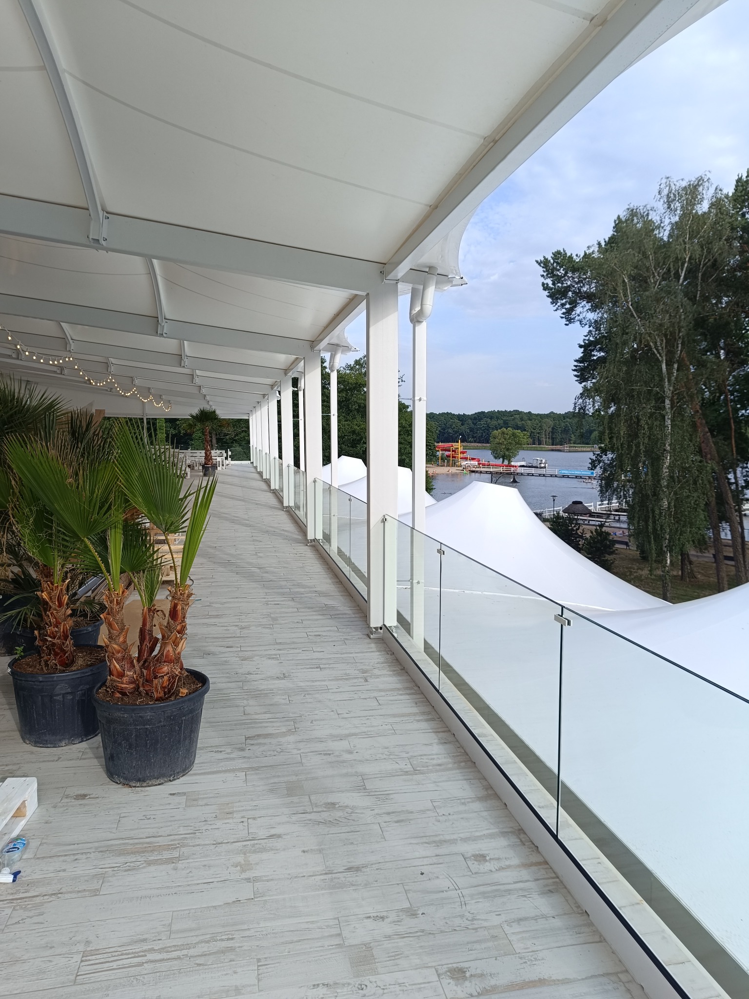Długi balkon z szarymi deskami podłogowymi, osłonięty białym zadaszeniem i zabezpieczony szklaną balustradą, z widokiem na jezioro i las w oddali. Na balkonie stoją donice z palmami.