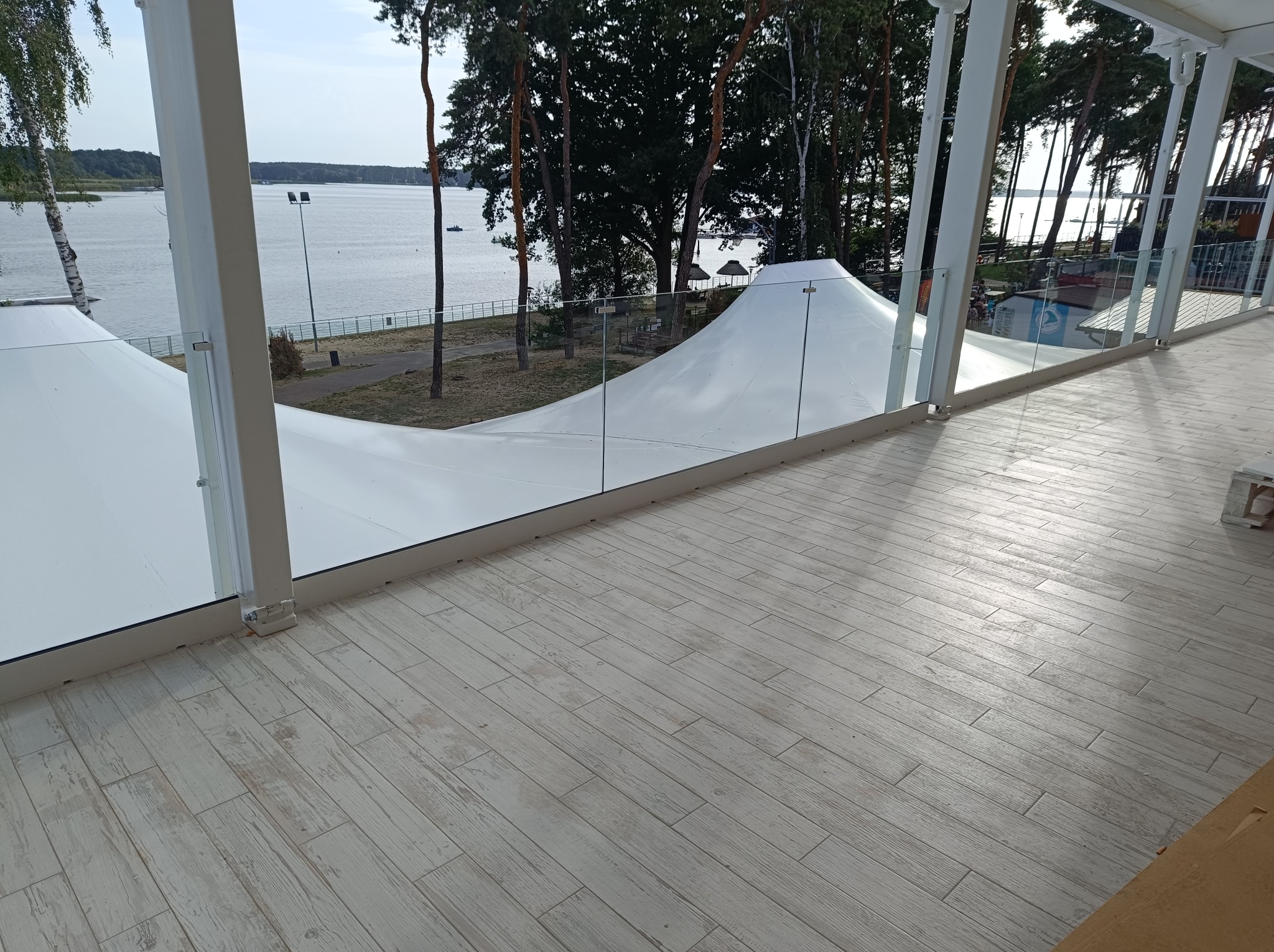 Szklana balustrada na tarasie z widokiem na jezioro i biały namiot, podłoga wyłożona płytkami imitującymi drewno.
