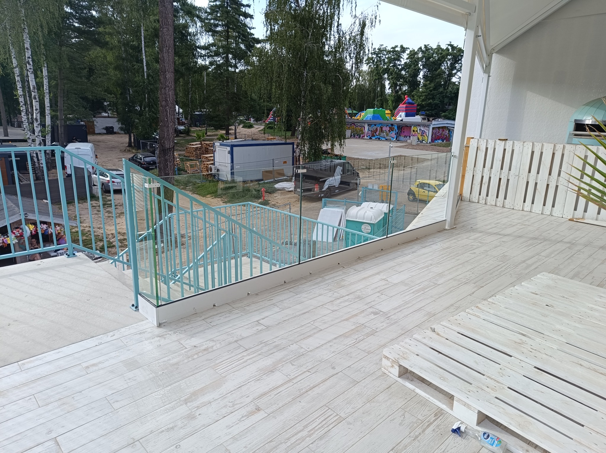 Turkusowa balustrada metalowa i szklana na tarasie z widokiem na plac zabaw i okoliczną zieleń.