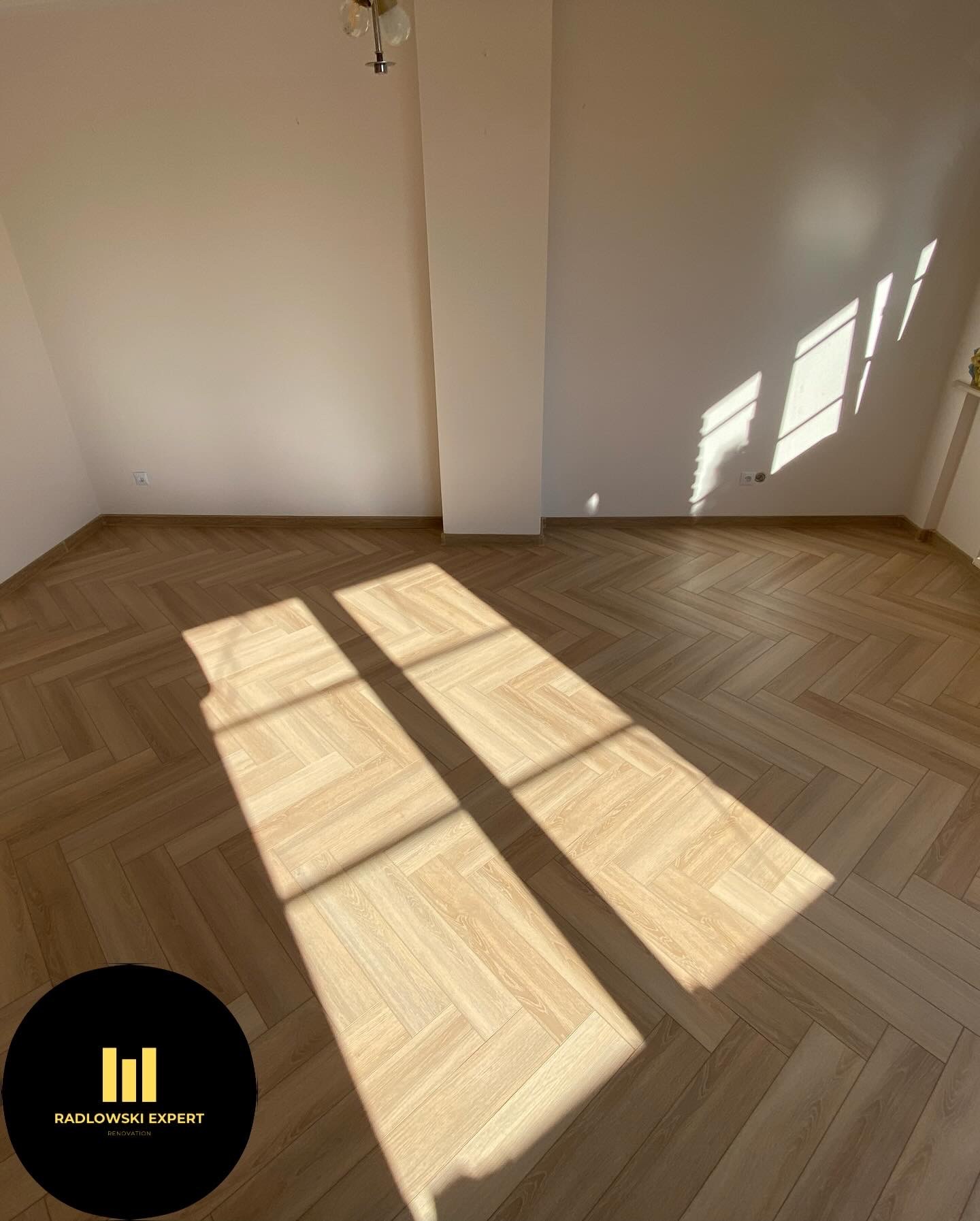 Jasne panele ułożone we wzór jodełki w pustym pokoju, oświetlone promieniami słońca wpadającymi przez okno, widoczne gniazdka elektryczne na ścianie, w rogu logo firmy remontowej.