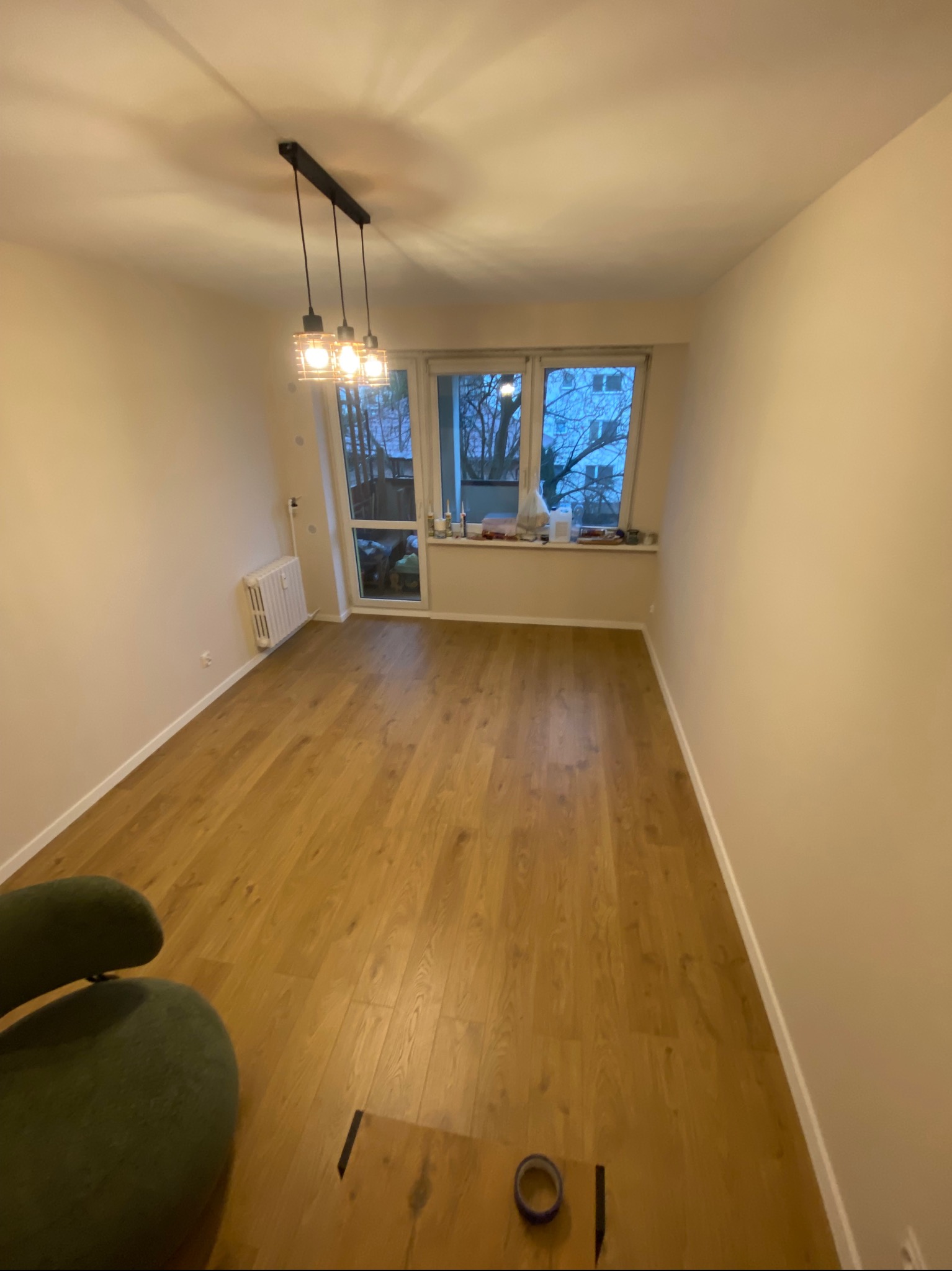 Pusty pokój po remoncie z drewnianą podłogą, świeżo pomalowanymi ścianami w odcieniach beżu, nowoczesną lampą wiszącą i widokiem na drzewa za oknem.