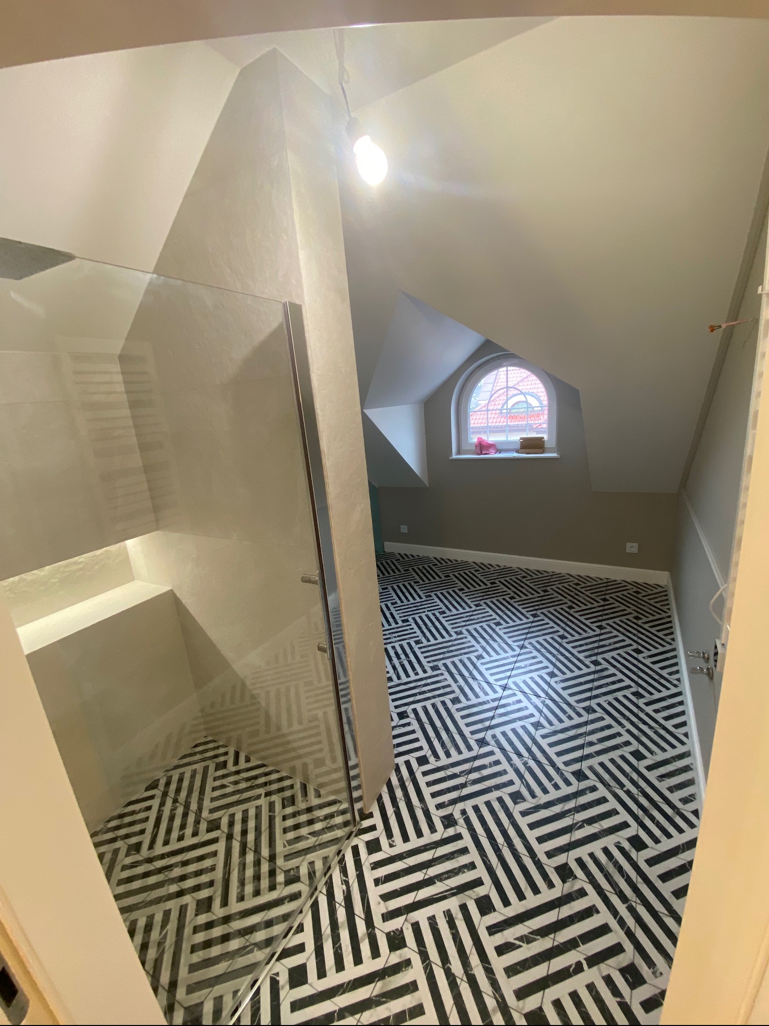 Łazienka na poddaszu z kabiną prysznicową typu walk-in i wzorzystą podłogą w czarno-białe pasy, widok zza uchylonych drzwi.