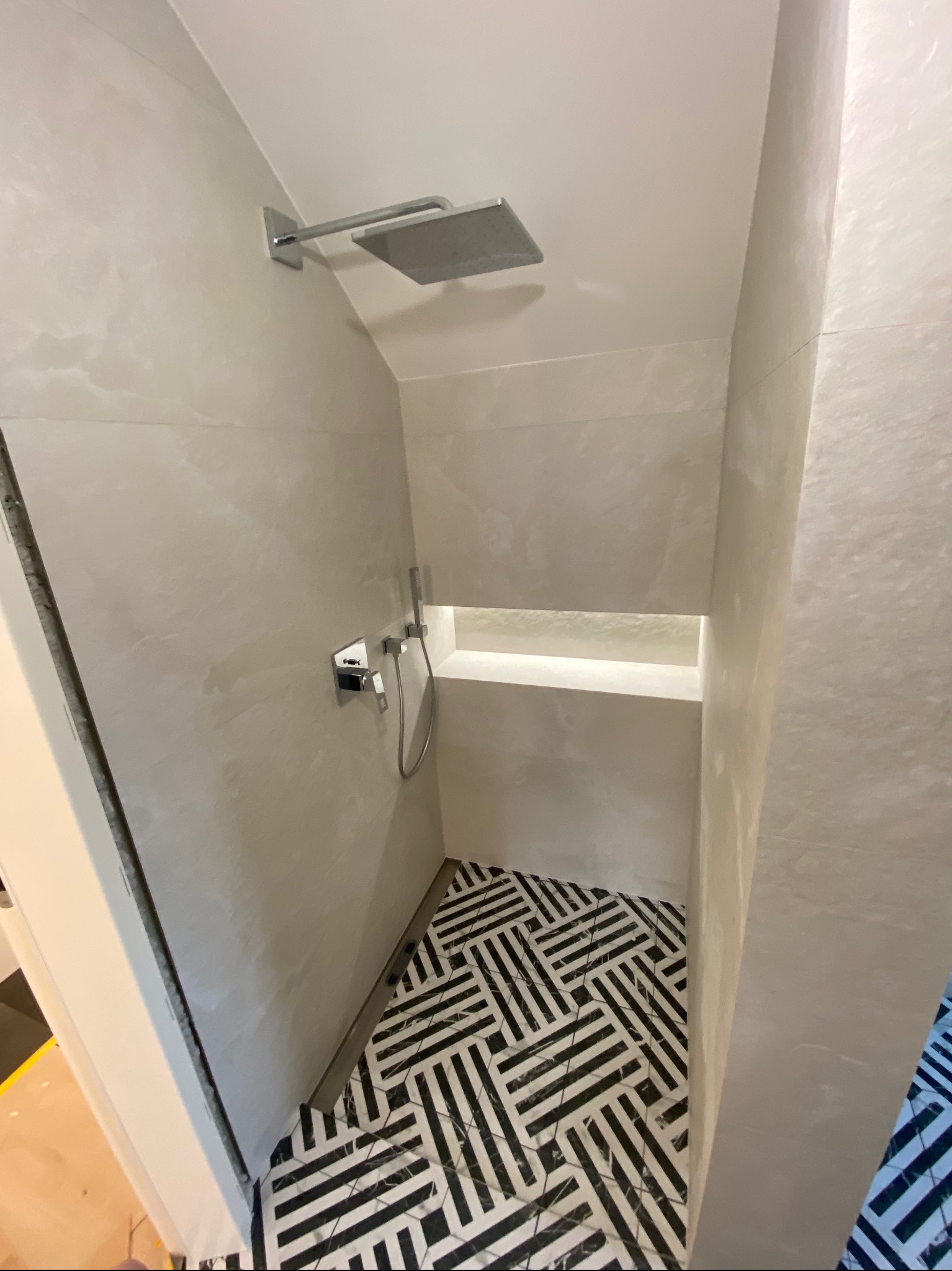 Nowoczesna kabina prysznicowa z deszczownicą i wnęką, wykończona jasnymi płytkami imitującymi kamień i posadzką w geometryczny wzór czarno-białych pasów.