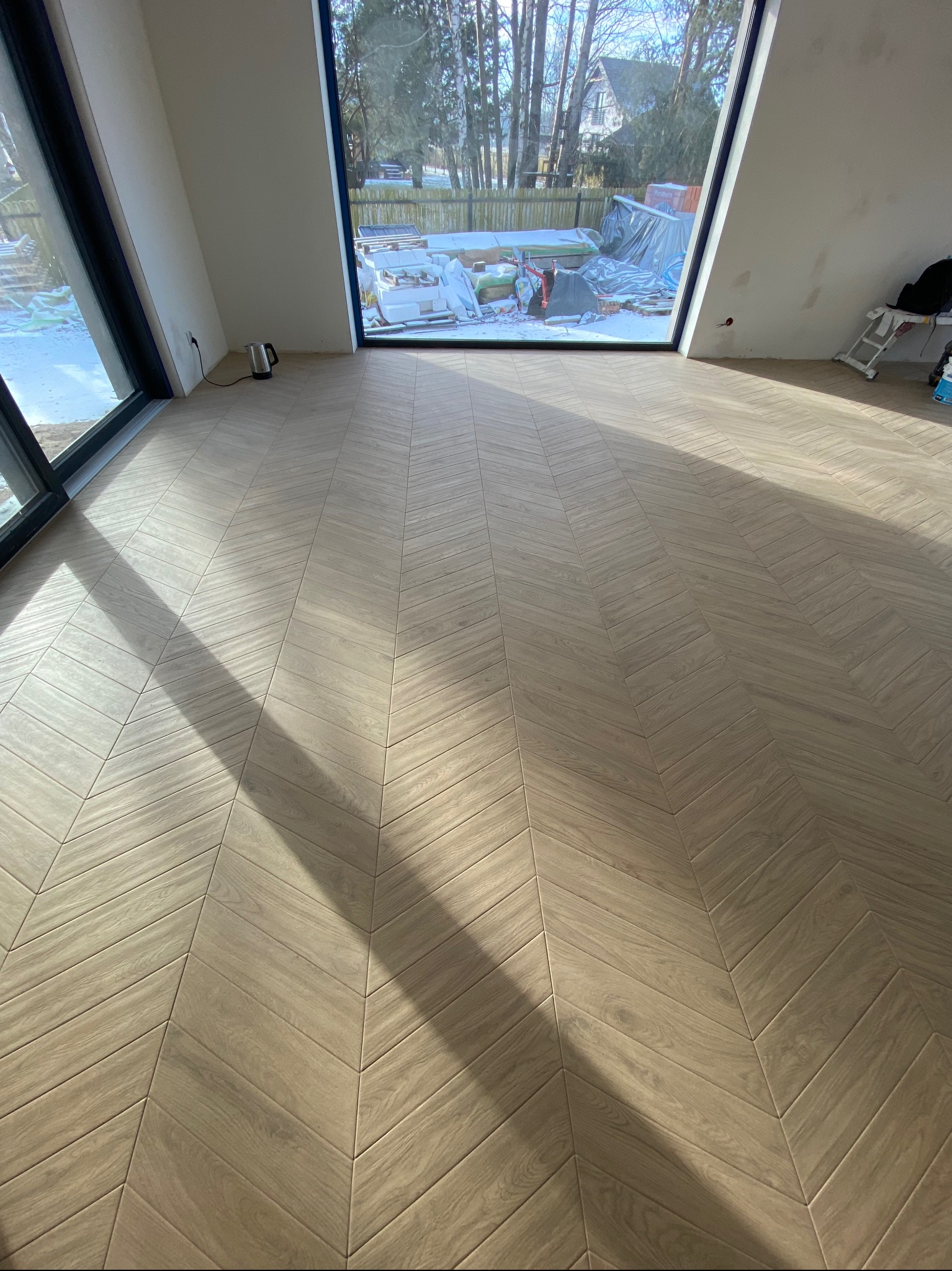 Ułożona podłoga z paneli w jodełkę w jasnym pomieszczeniu z dużymi oknami, widoczne refleksy światła słonecznego i fragmenty niezakończonych prac wykończeniowych.