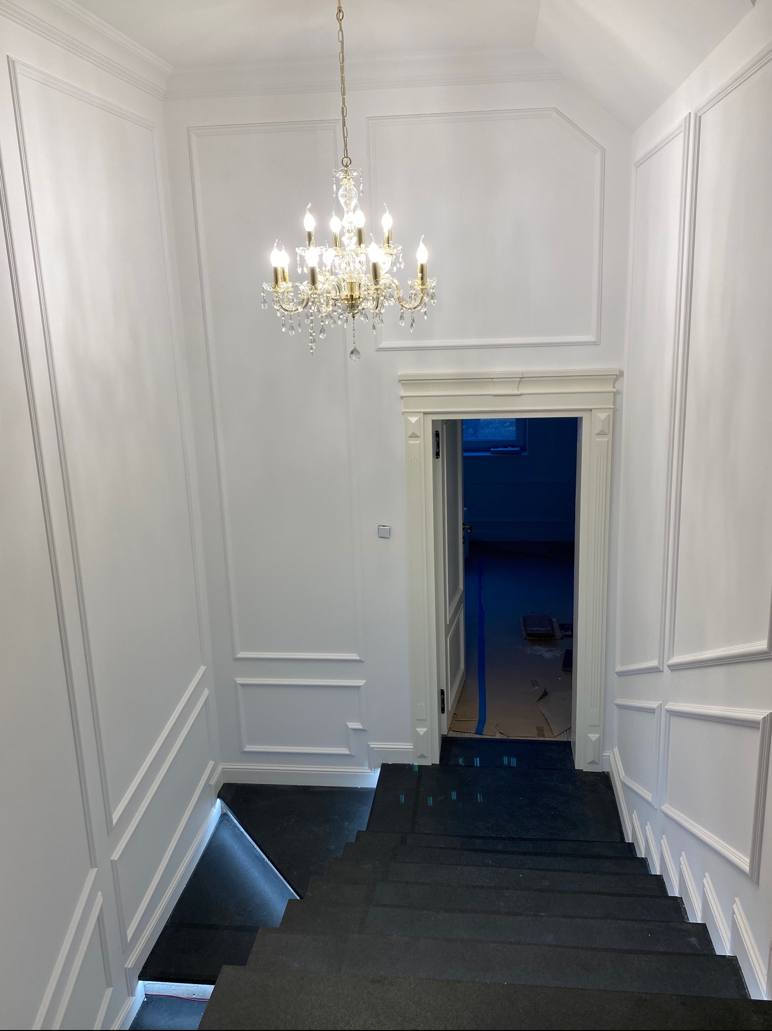 Klatka schodowa z czarnymi schodami i białymi ścianami z dekoracyjnymi listwami, kryształowy żyrandol pod sufitem, otwarte drzwi do ciemnego pomieszczenia.