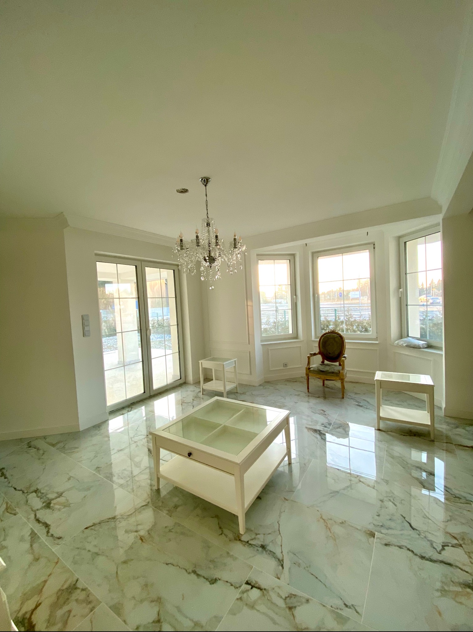 Jasne przestronne wnętrze z marmurową podłogą, kryształowym żyrandolem i widokiem z okna na zieleń.