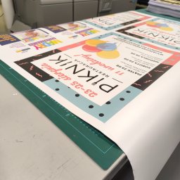 PS PRINT PIOTR STĘPNIEWSKI - Stos świeżo wydrukowanych plakatów reklamowych wydarzenia 'Piknik Restauracja 11 urodziny!' leżących na zielonej macie do cięcia, z widocznym fragmentem drukarki w tle.