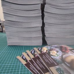 PS PRINT PIOTR STĘPNIEWSKI - Stosy świeżo wydrukowanych katalogów produktowych dla branży gastronomicznej, ułożone na zielonej macie do cięcia, z widocznymi detalami okładki i zawartości.