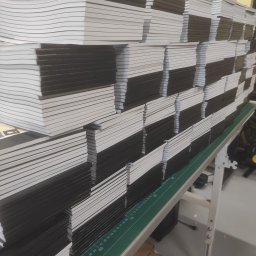 PS PRINT PIOTR STĘPNIEWSKI - Stosy świeżo wydrukowanych, czarnych zeszytów z białymi kartkami, ułożone na zielonej macie z podziałką centymetrową w drukarni.