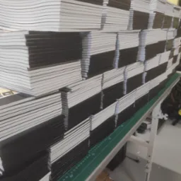 Stosy świeżo wydrukowanych, czarnych zeszytów z białymi kartkami, ułożone na zielonej macie z podziałką centymetrową w drukarni.