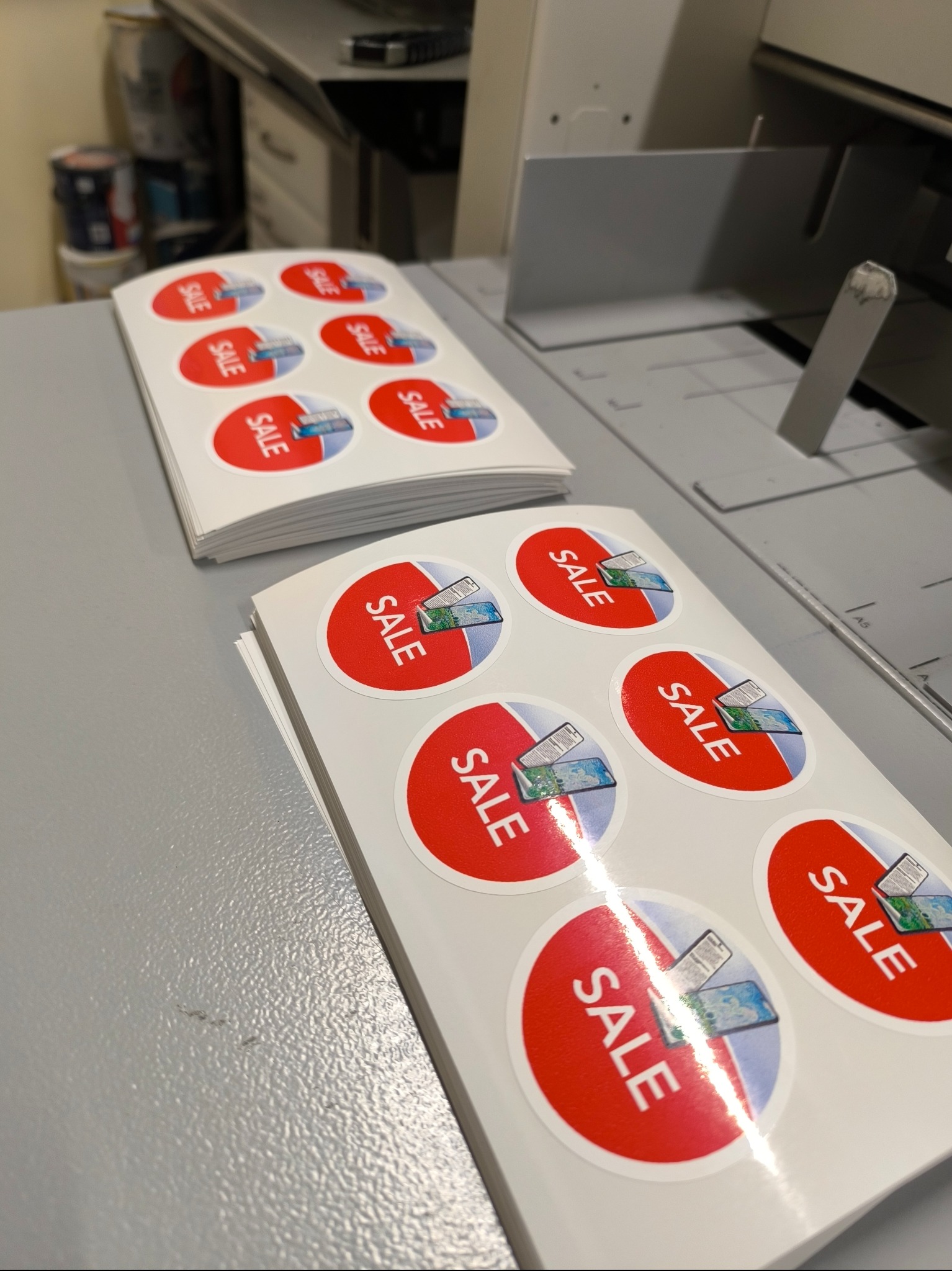 Stos naklejek okrągłych z czerwonym obramowaniem, napisem SALE i grafiką przedstawiającą dwa smartfony, ułożonych na szarym stole obok drukarki.