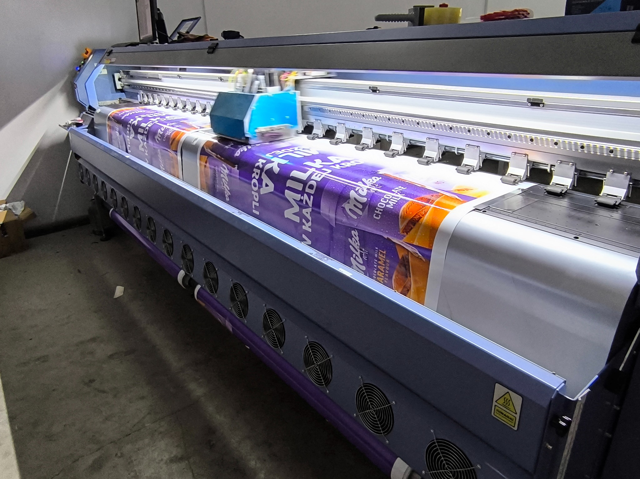 Szerokoformatowa drukarka w trakcie drukowania dużego baneru reklamowego z motywem czekolady Milka Caramel, widoczne detale mechanizmu drukującego i wentylatory chłodzące.