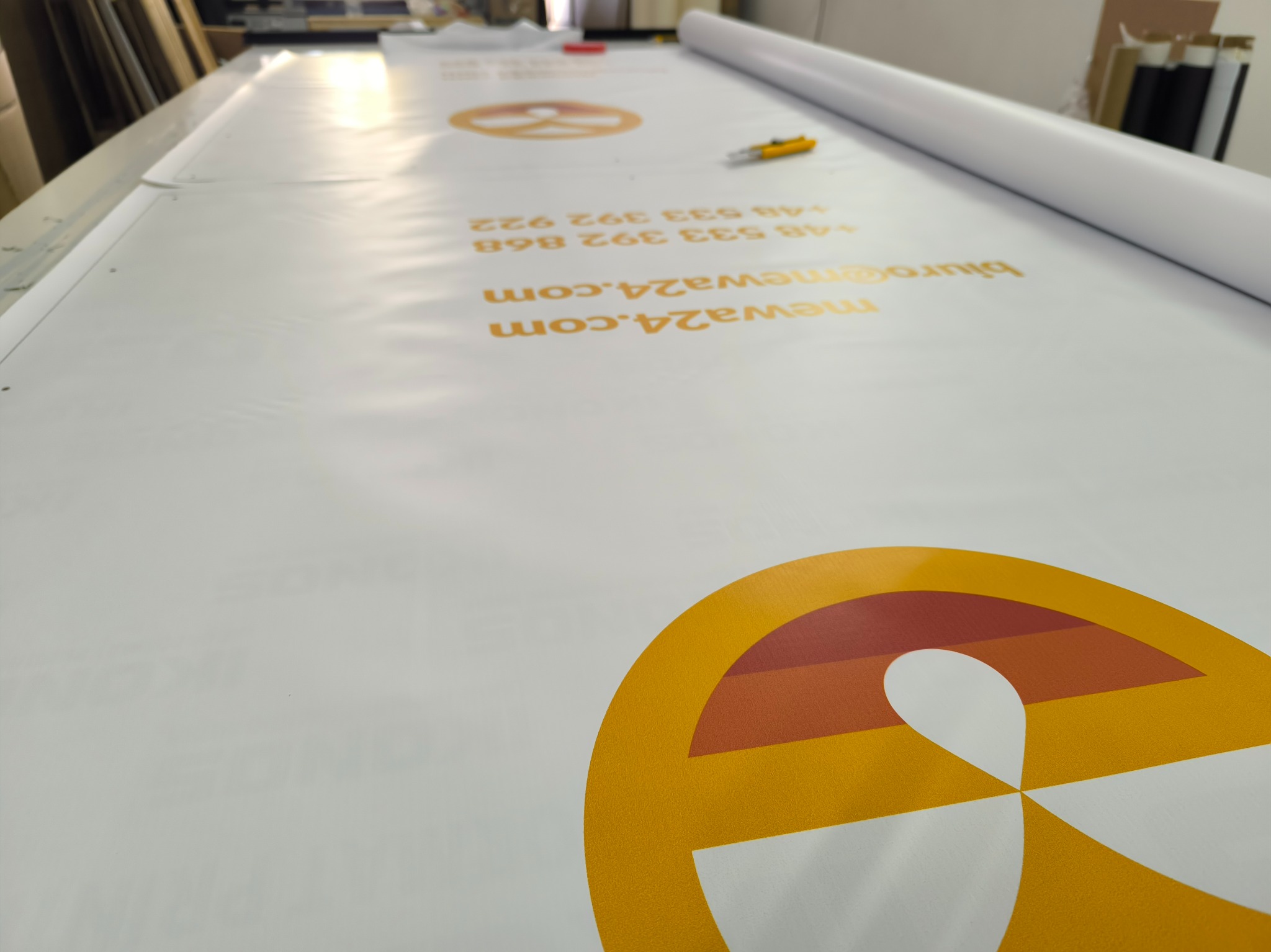 Długi baner z nadrukowanym pomarańczowym logo i danymi kontaktowymi leży na stole w drukarni, obok rolka niezadrukowanego materiału i nóż introligatorski.