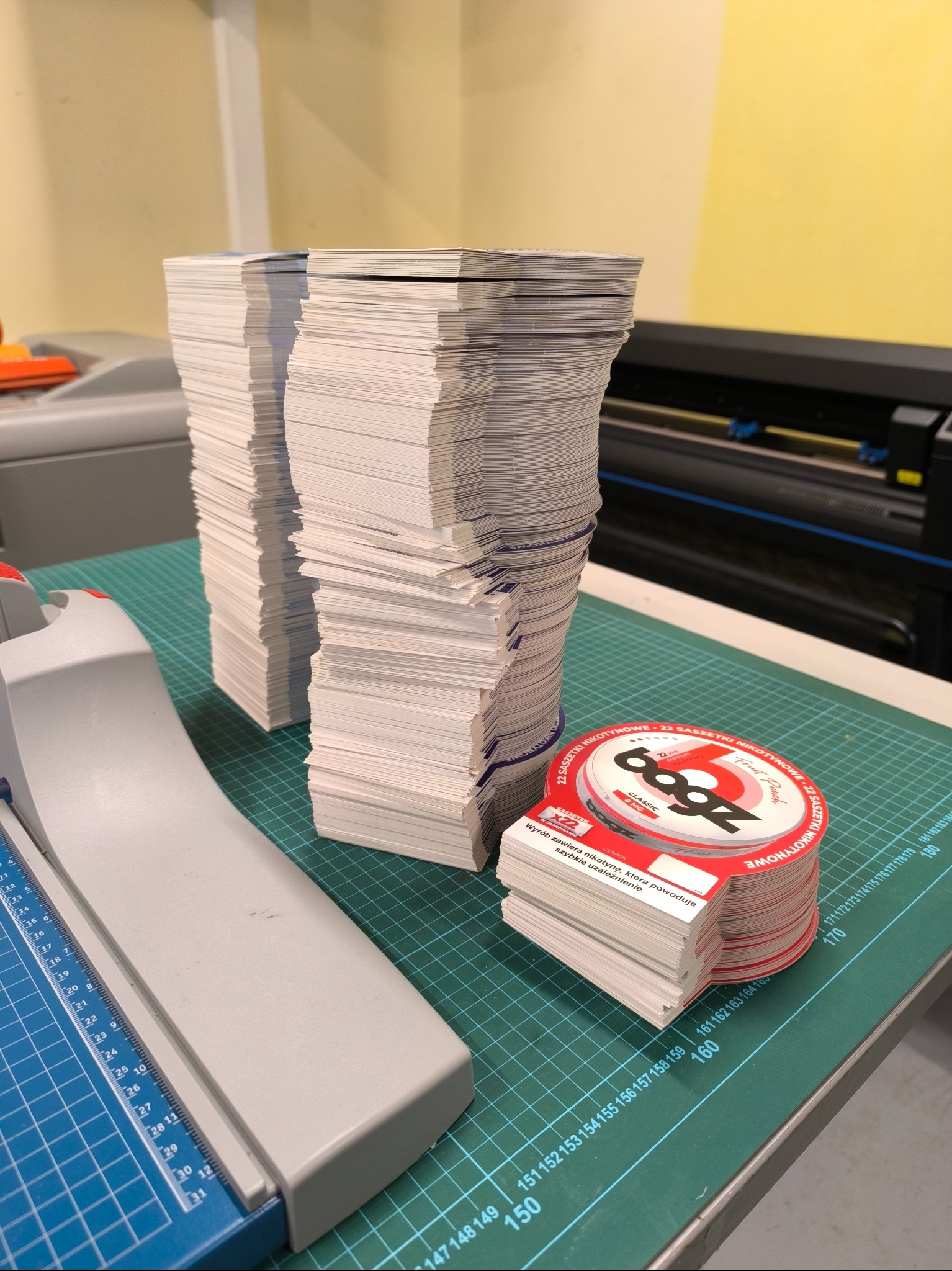 Stosy świeżo przyciętych, kwadratowych kartoników obok gilotyny do papieru i okrągłych naklejek z informacjami o produkcie na macie do cięcia.
