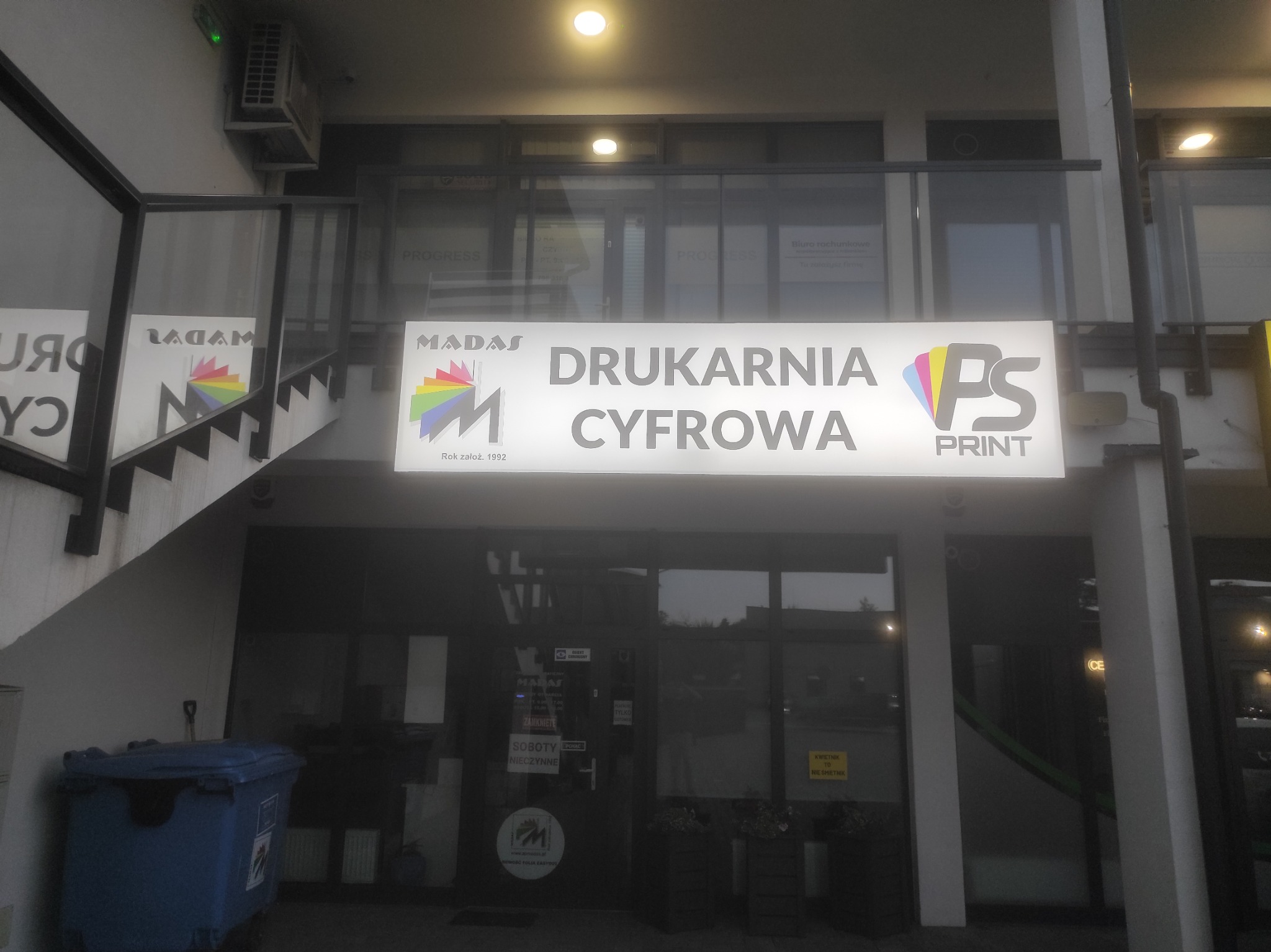 Fasada budynku z szyldem 'MADAS Drukarnia Cyfrowa PS PRINT', widoczny kosz na śmieci, schody i okna biurowe.