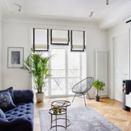 Elegancki salon w stylu paryskim z granatową sofą Chesterfield, parkietem w jodełkę, białymi ścianami i roletami rzymskimi z granatowym obszyciem.