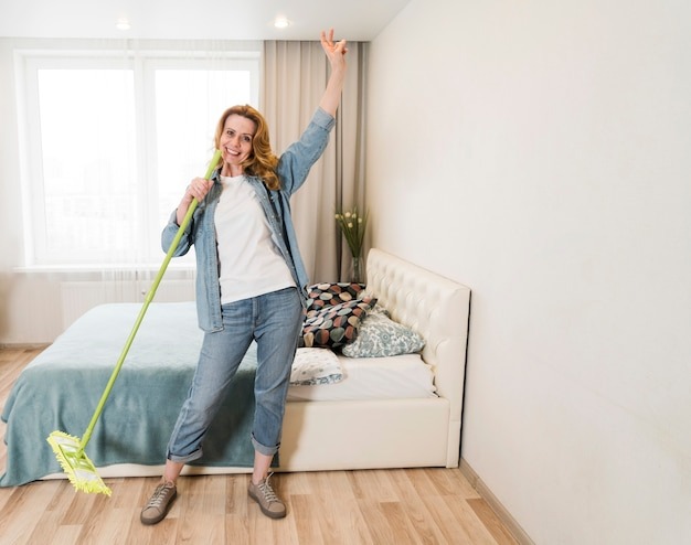 Uśmiechnięta kobieta w jeansowej koszuli, trzymająca zielony mop w sypialni, z podniesioną ręką w geście zwycięstwa, na tle jasnego pokoju z łóżkiem i oknem.