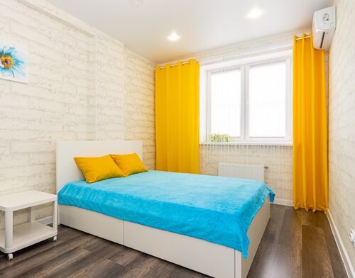 Jasna sypialnia z białymi ścianami imitującymi cegłę, turkusową narzutą na łóżku, żółtymi zasłonami, białymi meblami i klimatyzacją na ścianie.