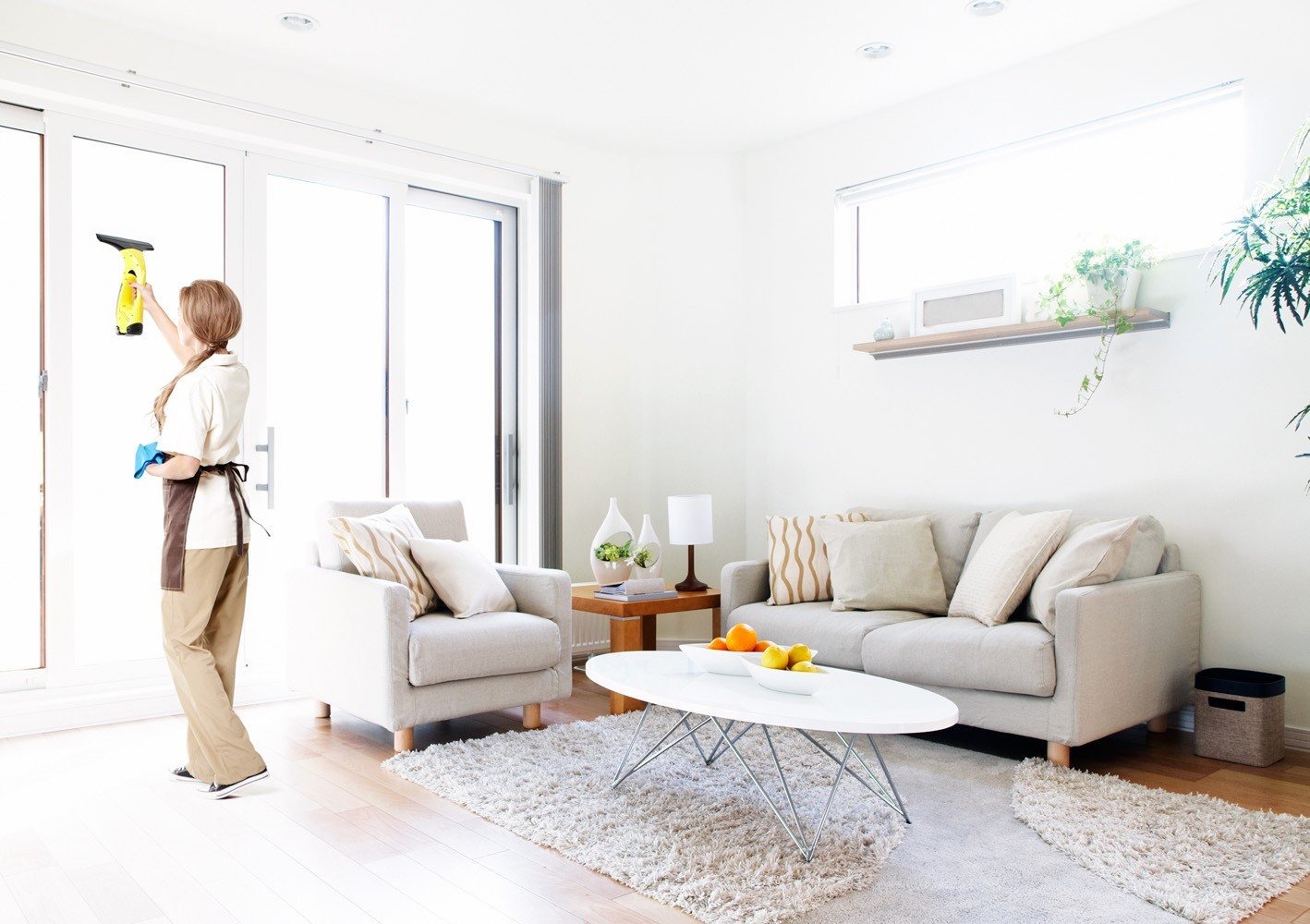 Kobieta w uniformie czyści szklane drzwi balkonowe w jasnym, nowocześnie urządzonym salonie, używając ściągaczki i niebieskiej ściereczki.